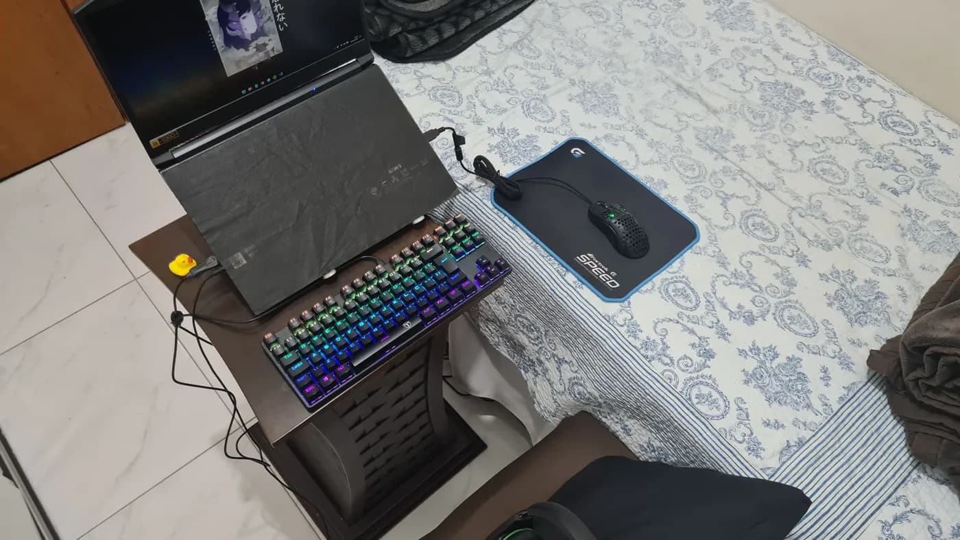 Notebook, teclado mecânico e mouse montados numa mesinha ao lado da cama: o setup dos estudos